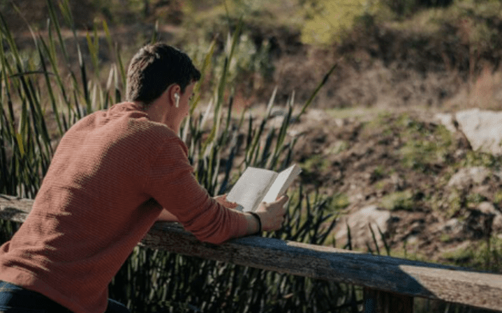 A man reading a book on top of a wooden fence.