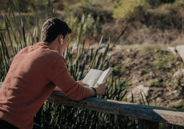 A man reading a book on top of a wooden fence.